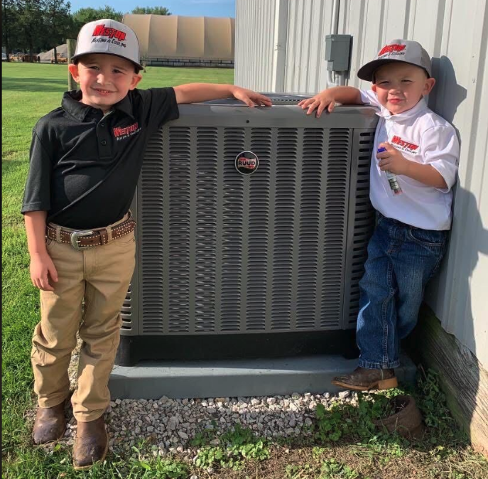 Two kids in Weston Heating & Cooling gear standing next to a Ruud outdoor AC unit